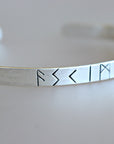 A silver bangle bracelet with a row of four Norse runes engraved on the front. The runes are F, S, K, and I.