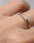 Two rose gold rings are shown on a finger. One ring is a simple band, and the other has a twisted rope design. The rings are delicate and appear to be made of a shiny metal.