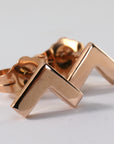 A pair of rose gold chevron stud earrings. The earrings are small and delicate, and they have a simple, modern design.