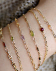 Five gold chain bracelets with different colored gemstones. The bracelets feature a delicate chain design and small, oval-shaped gemstones in shades of red, green, purple, and orange.
