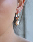 Rose gold hoop earrings with a large, off-white pearl charm dangling from one earring.