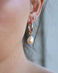 Rose gold hoop earrings with a large, off-white pearl charm dangling from one earring.