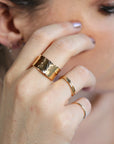 A close-up of a hand wearing three gold rings. The largest ring is a wide band with a hammered texture. The other two rings are thinner and more delicate.