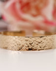 A gold filled cuff bracelet with a hammered texture. The bracelet has a wide band and an open design. The background is blurred, with a hint of pink and white.