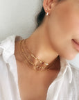 A close-up of a person wearing three gold necklaces. The necklaces are layered and feature different chain styles, including a delicate chain, a curb chain, and a chain with small, round charms. The person is also wearing a pair of small, gold stud earrings.