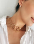A close-up of a person wearing three gold necklaces. The necklaces are layered and feature different chain styles, including a delicate chain, a curb chain, and a chain with small, round charms. The person is also wearing a pair of small, gold stud earrings.