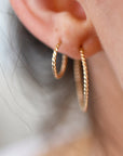 A pair of gold-toned hoop earrings with a twisted rope design. The earrings are worn in the earlobe.