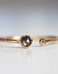 A gold ring with a single, round, grey diamond set in the center. The ring is open on one side and has a textured band.