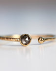 A gold ring with a single, round, grey diamond set in the center. The ring is open on one side and has a textured band.