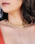 A gold chain necklace with a simple, linked design. The necklace is worn by a person with dark hair and a red top.