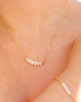A delicate gold chain necklace with a row of small white opal beads. The necklace is worn by a person, and a matching bracelet with white opal beads is also visible on the wrist.