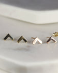 Two pairs of gold chevron stud earrings. The earrings are arranged in a row on a white surface. The earrings are small and delicate. The earrings are made of solid gold and have a simple design for daily use