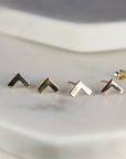 Two pairs of gold chevron stud earrings. The earrings are arranged in a row on a white surface. The earrings are small and delicate. The earrings are made of solid gold and have a simple design for daily use