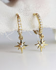 A pair of gold hoop earrings with a dangling star charm. Each hoop is encrusted with small, clear stones and the star charm has a single, larger clear stone in the center.