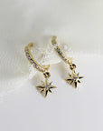 Two gold hoop earrings with a dangling star charm. Each hoop is lined with small clear stones and each star charm has a single clear stone in the center.