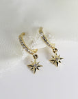 Two gold hoop earrings with a dangling star charm. Each hoop is lined with small clear stones and each star charm has a single clear stone in the center.