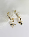 Two gold hoop earrings with a dangling star charm. Each hoop is lined with small clear stones and each star charm has a single clear stone in the center.