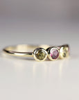 May include: A gold band ring with four round gemstones. The gemstones are green, pink, green, and green. The ring is on a white surface.