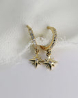 Two gold hoop earrings with a dangling star charm. The hoops are lined with small clear diamond stones and the stars have a single clear diamond stone in the center.