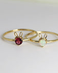 Two gold rings with bunny ear accents. One ring features a pink tourmaline gemstone and the other features a white opal.