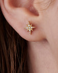 A gold starburst stud earring with small, clear diamonds in the center. The earring is worn on an ear, and the design features a detailed, textured surface. The starburst design is a classic and elegant jewelry piece.