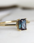 A gold ring with a dark blue, emerald-cut gemstone. The ring is simple and elegant.