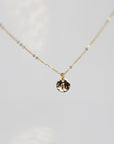A gold chain necklace with a small, round pendant featuring the letter "a" in script font on a hammered finish disc.
