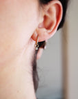 A gold hoop earring with a small diamond dangling from it. The earring is worn in the left ear of a person with dark hair.