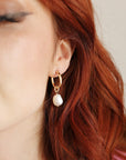 Gold hoop earrings with a dangling pearl charm. The earrings are worn by a person with red hair.