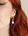 Gold hoop earrings with a dangling pearl charm. The earrings are worn by a person with red hair.