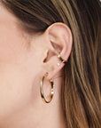 Gold hoop earrings and ear cuff jewelry. The hoop earrings are large and round. The ear cuff is a thin band that wraps around the upper part of the ear.