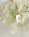 A gold necklace with a round pendant featuring a small engraved cactus design. The necklace is displayed on a white background with white flowers.