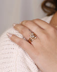 A gold ring with a large round center stone and smaller stones surrounding it. The ring is set on a thin gold band. The ring is on a finger with a gold band on the finger next to it.
