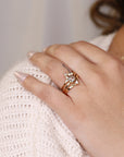 A gold ring with a large round center stone and smaller stones surrounding it. The ring is set on a thin gold band. The ring is on a finger with a gold band on the finger next to it.