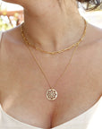 A gold chain necklace with a delicate pendant featuring a circular design with intricate cutouts. The necklace is layered over a thicker gold chain.