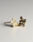 A pair of gold butterfly stud earrings. Each earring features a detailed butterfly design with etched wings and body. The earrings are displayed on a white surface, with one earring in focus and the other slightly blurred.