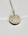A gold pendant necklace with a round disc charm. The disc has a constellation design with small diamonds set into the metal.