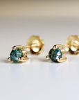 Close-up of a pair of 14k gold stud earrings featuring round, faceted teal gemstones. Each earring has four small gold accents surrounding the gemstone. The earrings are set against a blurred, light background. The image highlights the earrings&