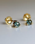 Close-up of a pair of gold green sapphire stud earrings, each featuring a faceted, dark teal gemstone. The gemstones are round and securely set in four-prong settings. The earrings have a simple, elegant design, suitable for everyday wear or special occasions. The gold is a warm, yellowish tone, and the gemstones have a subtle sparkle. The earrings are shown against a light gray background.