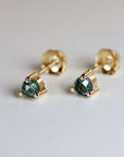 Close-up of a pair of gold stud earrings featuring faceted, dark-green gemstones. Each earring has a small, round gemstone securely set in a four-prong gold setting. The earrings have a simple, elegant design, suitable for everyday wear or special occasions. The gold posts are visible, indicating they are designed for pierced ears. The gemstones appear to have a subtle sparkle, suggesting a possible high-quality material.