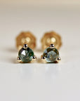 Close-up of a pair of gold stud earrings featuring round, faceted green gemstones. Each earring has three small gold balls at the base of the setting. The gemstones have a dark, slightly bluish-green hue and exhibit sparkle and depth. The earrings are elegantly simple and showcase the natural beauty of the stones. The image highlights the delicate craftsmanship and the subtle color variations within the gemstones.
