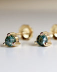 Close-up view of a pair of gold stud earrings featuring faceted, dark teal gemstones. Each earring has four small gold prongs securing the gemstone. The gemstones have a subtle sparkle and appear to be round or slightly faceted. The sapphire earrings are set against a light-colored, blurred background.