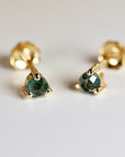 Close-up view of a pair of gold stud earrings. Each earring features a small, faceted, dark teal gemstone securely set in a gold setting. The earrings have a simple, elegant design, suitable for everyday wear or special occasions. The gemstones display subtle variations in color and sparkle. The gold posts are visible, indicating a standard stud earring design. The image highlights the delicate craftsmanship and the rich color of the gemstones.