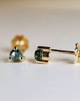 Close-up view of a pair of gold stud earrings featuring round, dark teal gemstones. Each earring has a small, three-prong setting and a post backing. The earrings are displayed against a bright, off-white background. The stones appear faceted and have a subtle sparkle. The gold is a warm, yellowish tone. The overall style is simple and elegant, suitable for everyday wear or special occasions. The image highlights the color and cut of the gemstones and the delicate design of the earrings.