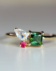 A gold ring with a green, square-shaped gemstone, a white pear-shaped gemstone, and a small red gemstone.
