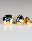 Two gold stud earrings with a light blue gemstone center. The earrings have a circular design with a polished finish. The gemstones are round and set in a bezel setting. The earrings are displayed on a white surface.
