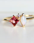 A gold ring featuring a square, pink gemstone, a white marquise-cut stone, and a small, round pink stone. The ring has a delicate band and is set against a white background. The gemstones are the focal point of the ring.