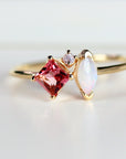 A gold ring featuring a square, pink gemstone, a white marquise-cut stone, and a small, round pink stone. The ring has a delicate band and is set against a white background. The gemstones are the focal point of the ring.