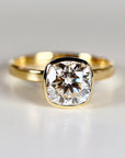 Cushion cut moissanite solitaire ring set in a 14k gold bezel, displayed on a white background as modern handcrafted engagement jewelry.