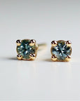 A pair of stud earrings featuring round, light blue sapphires. Each earring has a four-prong setting with small, rounded accents. The earrings are displayed against a white background.
